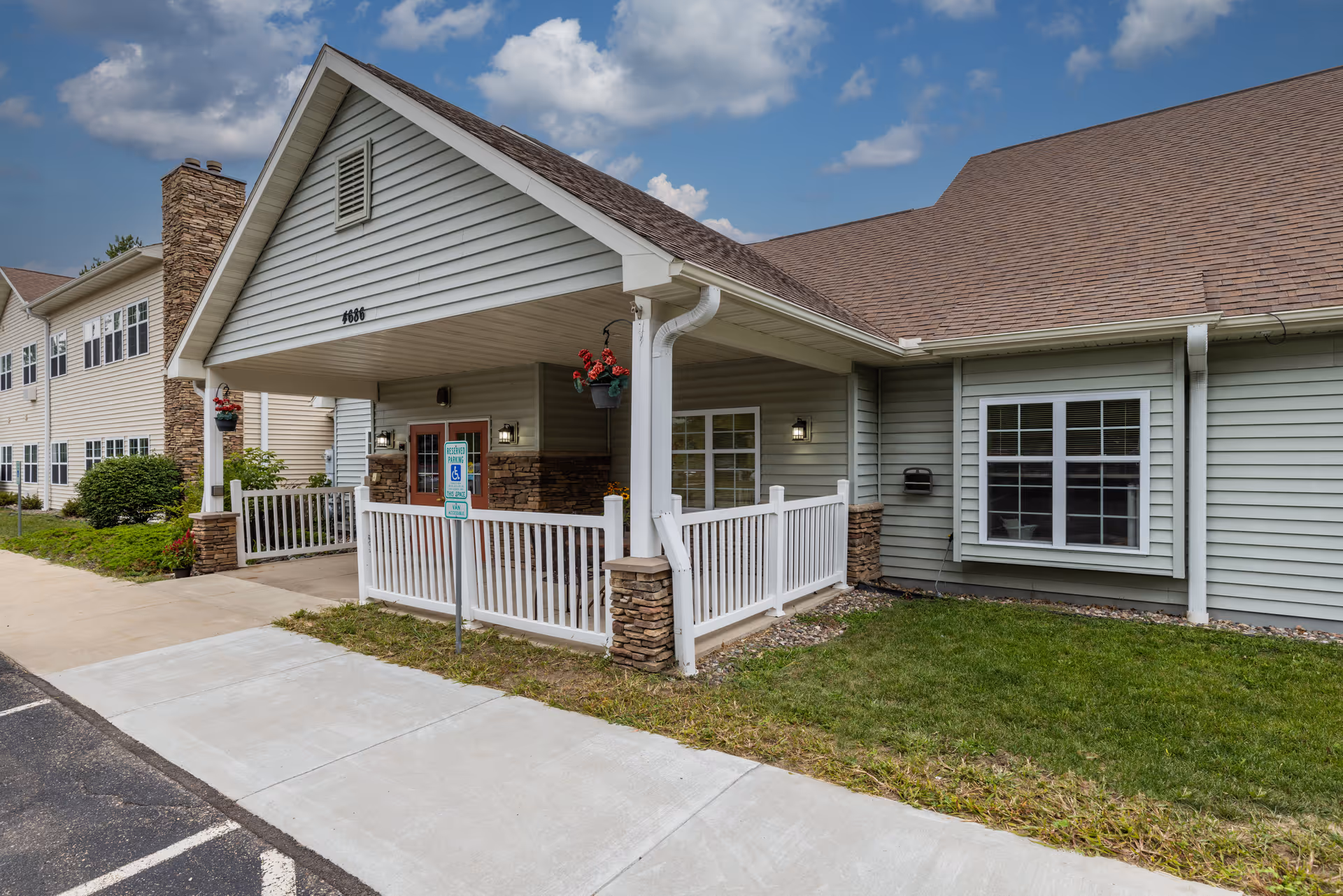 Covered front entrance of a senior living building with white railings, hanging flower baskets, and a paved walkway.