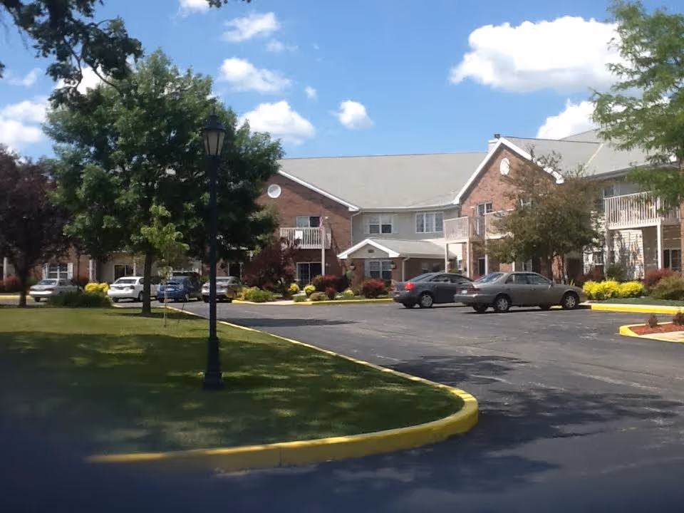 Front view of a two-story brick retirement apartment building with cars parked in a driveway, trees, and a blue sky.