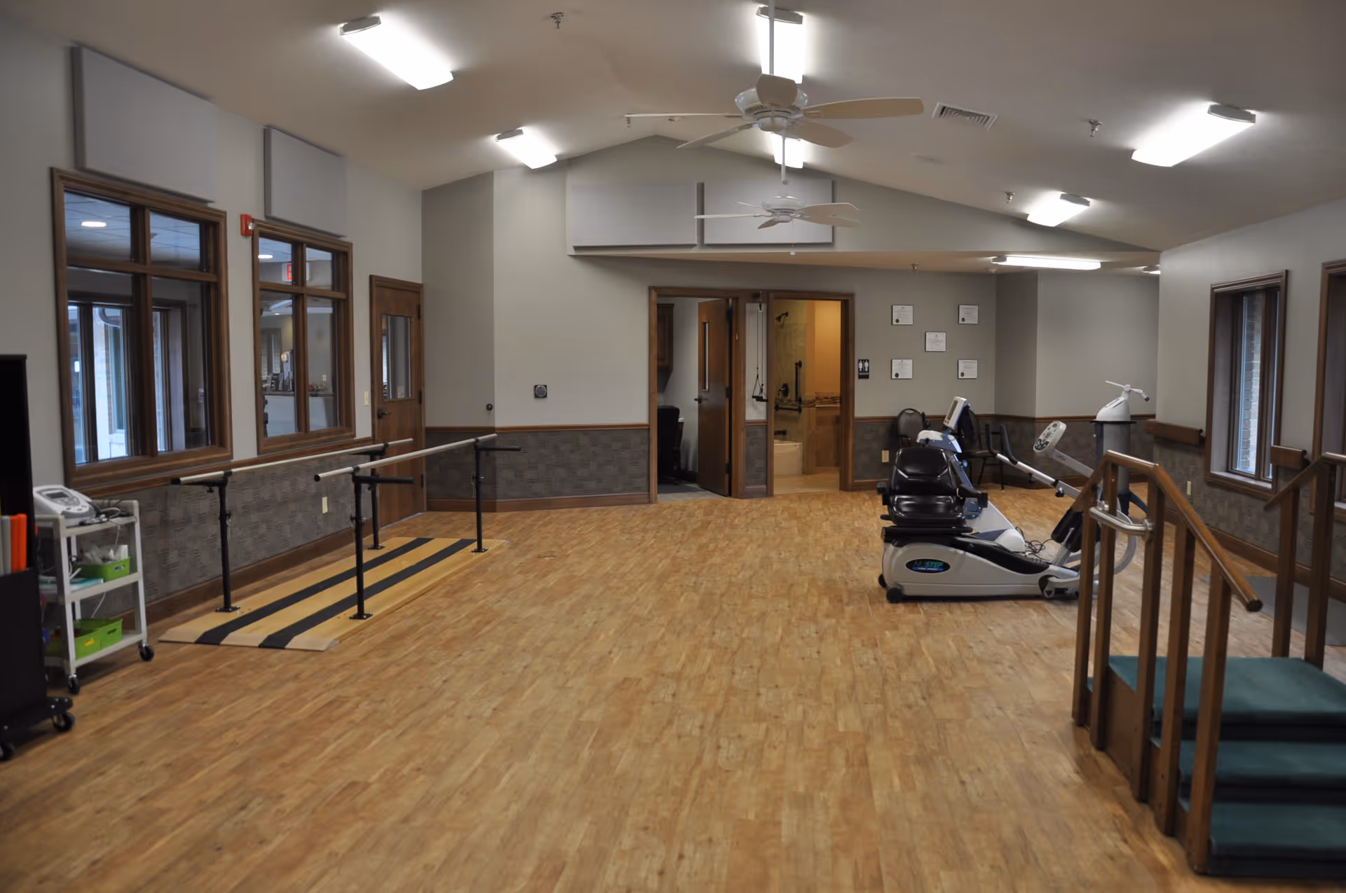 Spacious rehabilitation and exercise room with parallel bars, recumbent exercise bikes, handrails and a small step platform.