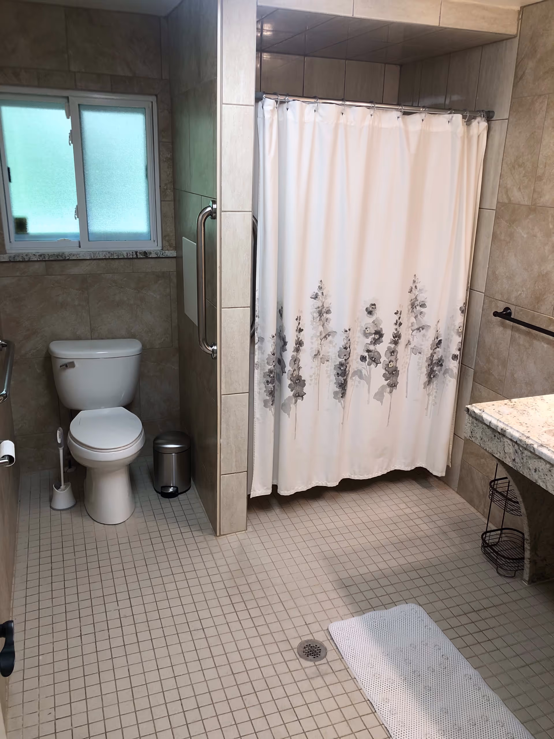 A bathroom with beige tiled walls and floor, featuring a white toilet with a toilet brush and small trash can beside it, a shower area with a white curtain decorated with gray floral patterns, a granite countertop with a black metal shelf underneath, and a frosted window providing natural light.