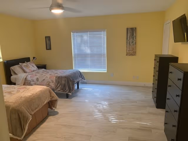 Bright yellow-walled bedroom with two twin beds, multiple dressers, a wall-mounted TV, and a window with blinds.