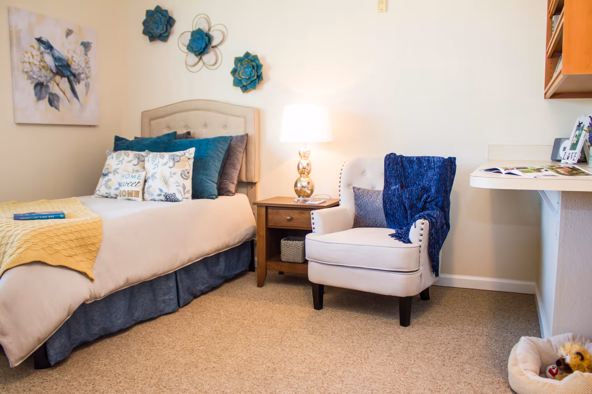 A cozy bedroom with a single bed adorned with multiple pillows and a yellow throw blanket. Next to the bed is a wooden nightstand with a decorative lamp. A comfortable armchair with a blue throw blanket and a pillow is placed beside the nightstand. The walls are decorated with floral artwork and blue flower wall hangings. The room has beige carpeting and a small pet bed with a toy in the corner.