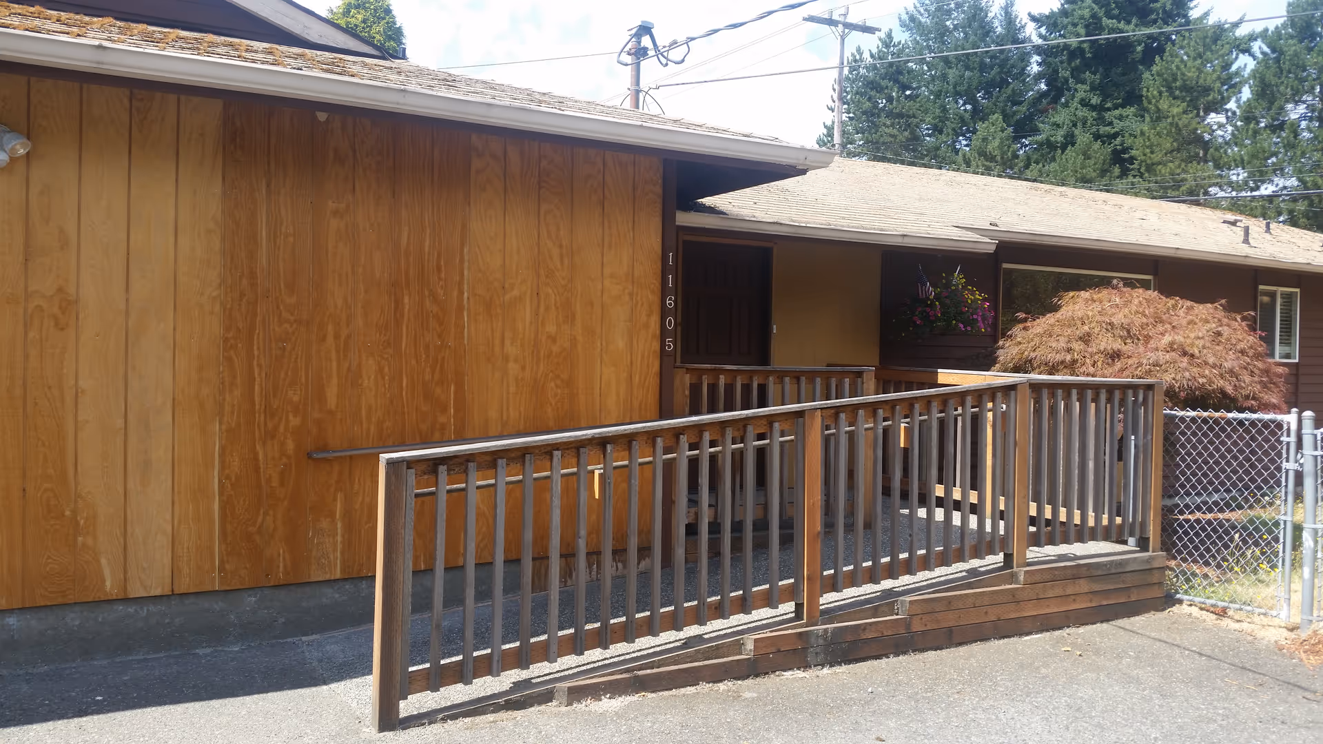 Exterior view of a single-story building with wooden panel siding and a sloped roof. There is a wooden ramp with railings leading to a door, and a small fenced area with a bush and hanging flowers near the entrance. Trees and power lines are visible in the background.