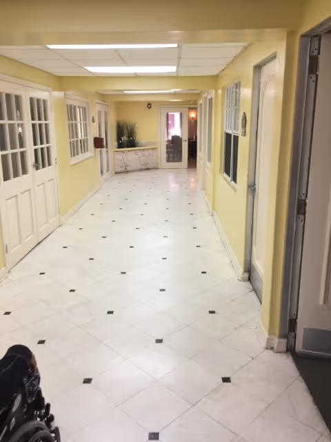 Long pale-yellow interior hallway with white tiled floor, doors and windows along the sides and a wheelchair partially visible in the foreground.