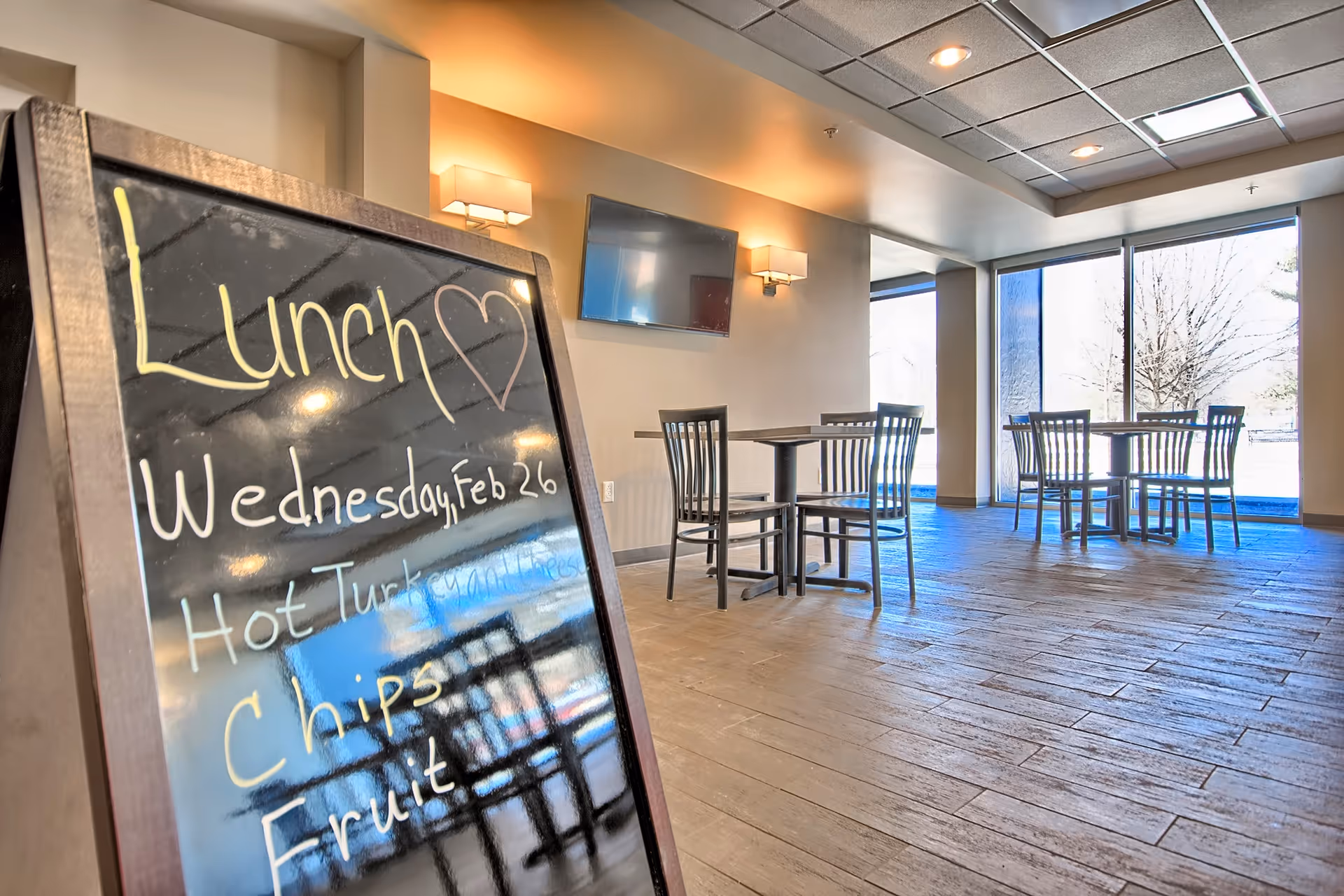 Dining room with a chalkboard lunch menu in the foreground and tables and chairs by large windows.