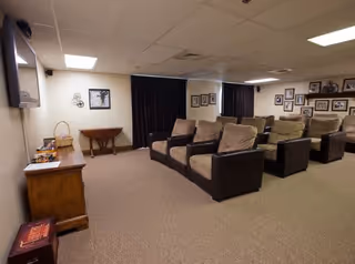A cozy common area with multiple cushioned armchairs arranged in rows facing a wall-mounted television. The room has beige carpeting, cream-colored walls, and a ceiling with recessed lighting. There are framed pictures on the walls and a wooden side table with some items on it.