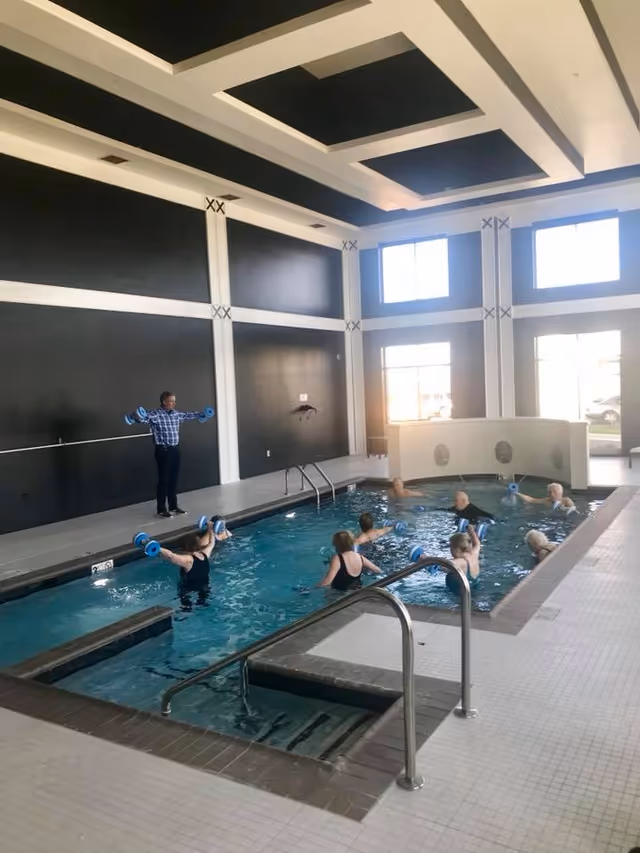 Indoor swimming pool with several elderly people participating in a water exercise class using blue foam dumbbells. An instructor stands at the poolside demonstrating the exercise. The room has high ceilings with black and white geometric patterns and large windows letting in natural light.