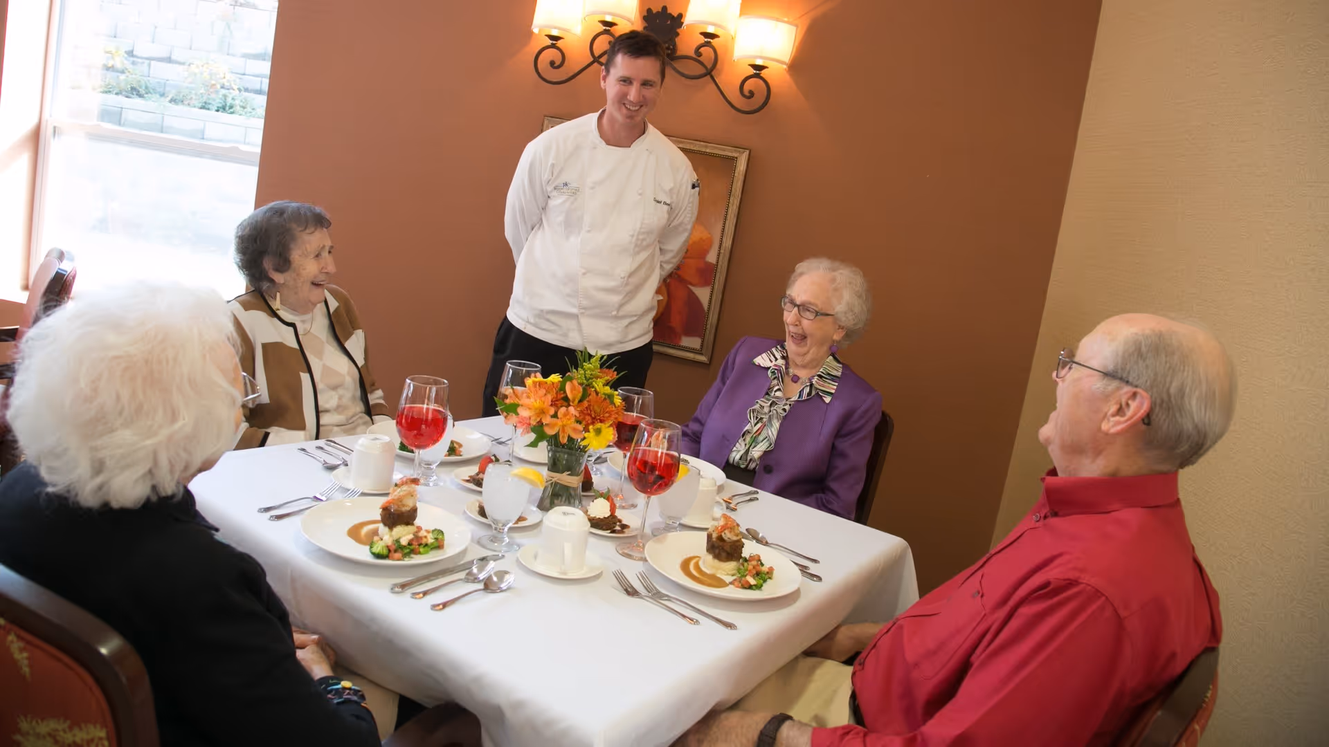 A chef stands and smiles at a dining table where four elderly people are seated, enjoying a meal together in a warmly lit room with a floral centerpiece on the table.