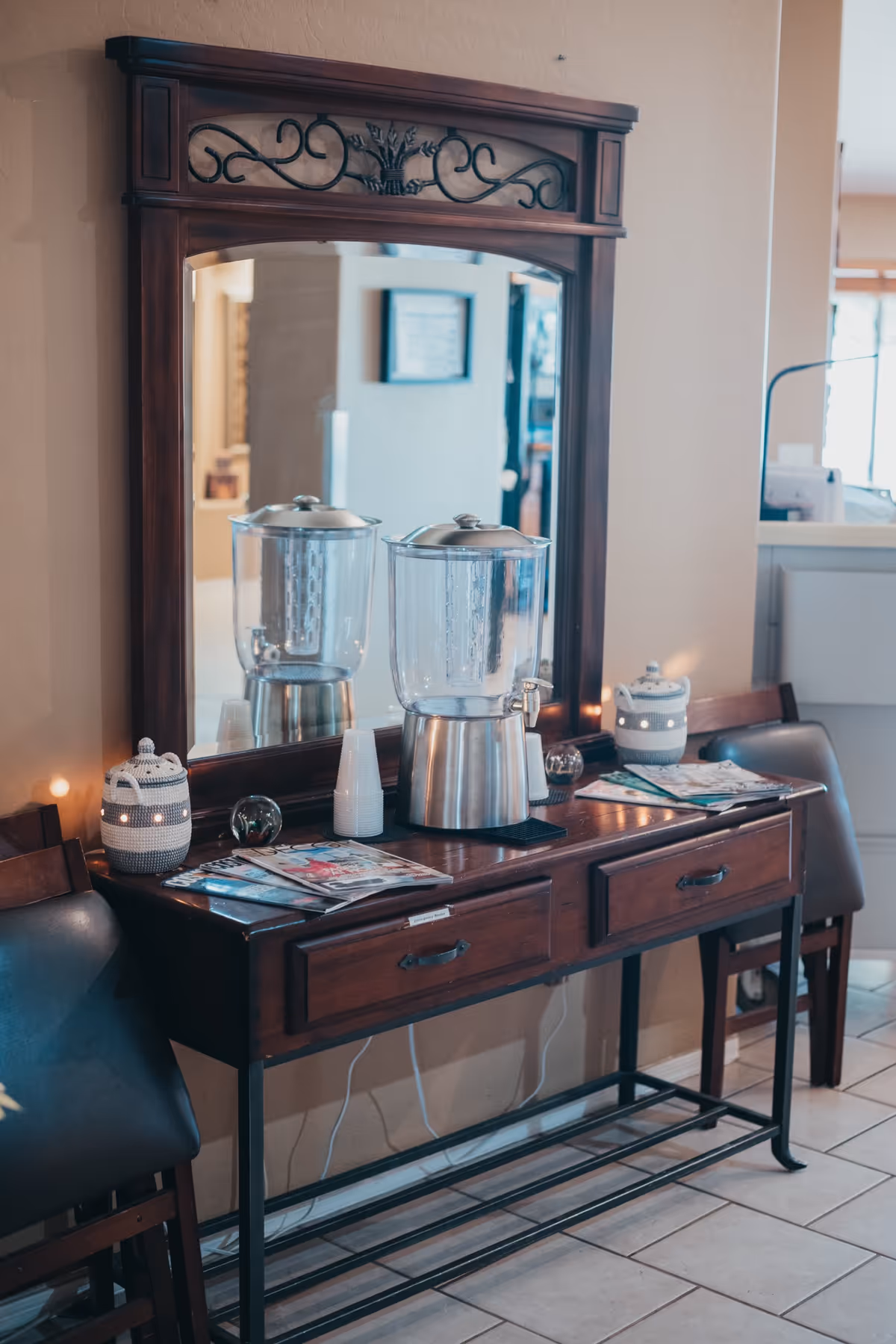A wooden console table with two drawers and a large decorative mirror above it. On the table, there is a water dispenser, a stack of disposable cups, some magazines, and two small decorative containers. Two chairs are positioned on either side of the table, and the setting appears to be inside a well-lit room with tiled flooring.