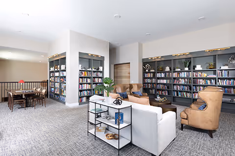 A bright and spacious common area with bookshelves filled with books along the walls, a white sofa, two tan leather armchairs, a small black metal and glass shelving unit, and a wooden table with chairs in the background. The room has carpeted floors and neutral-colored walls.