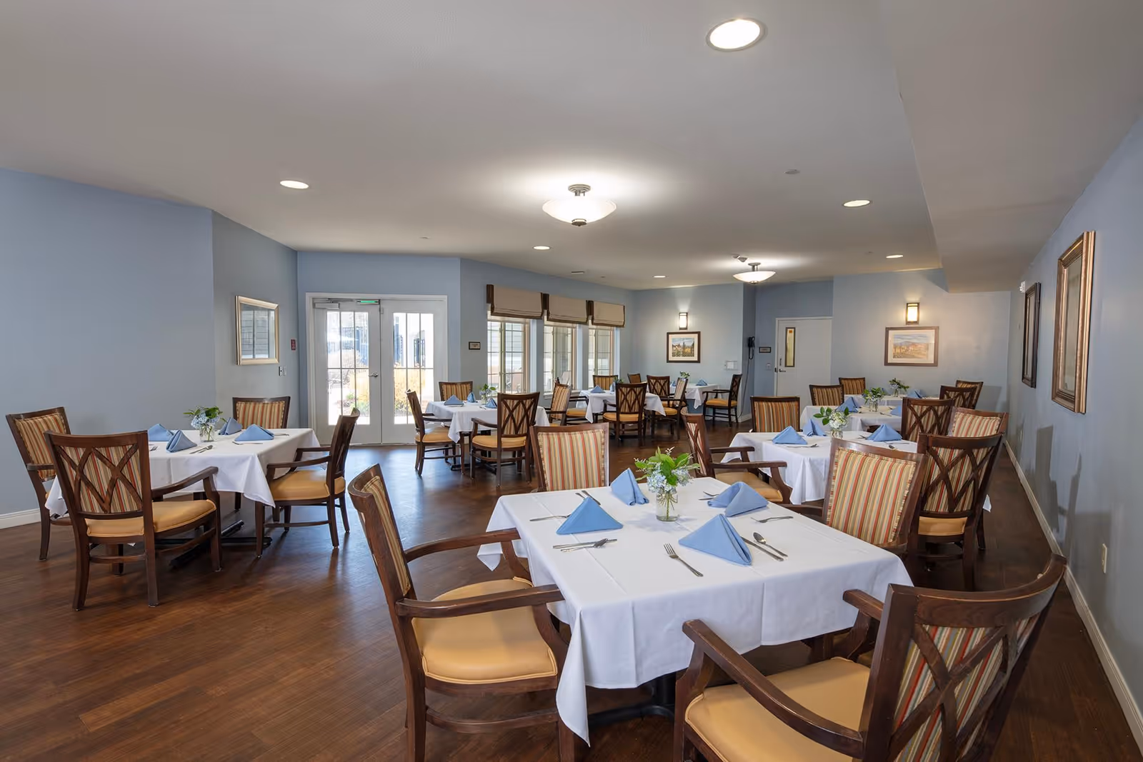 A dining room in a senior living facility with multiple tables covered in white tablecloths, each set with blue folded napkins, silverware, and small floral centerpieces. The room has wooden chairs with cushioned seats and striped backs, light blue walls, framed artwork, and large windows letting in natural light.