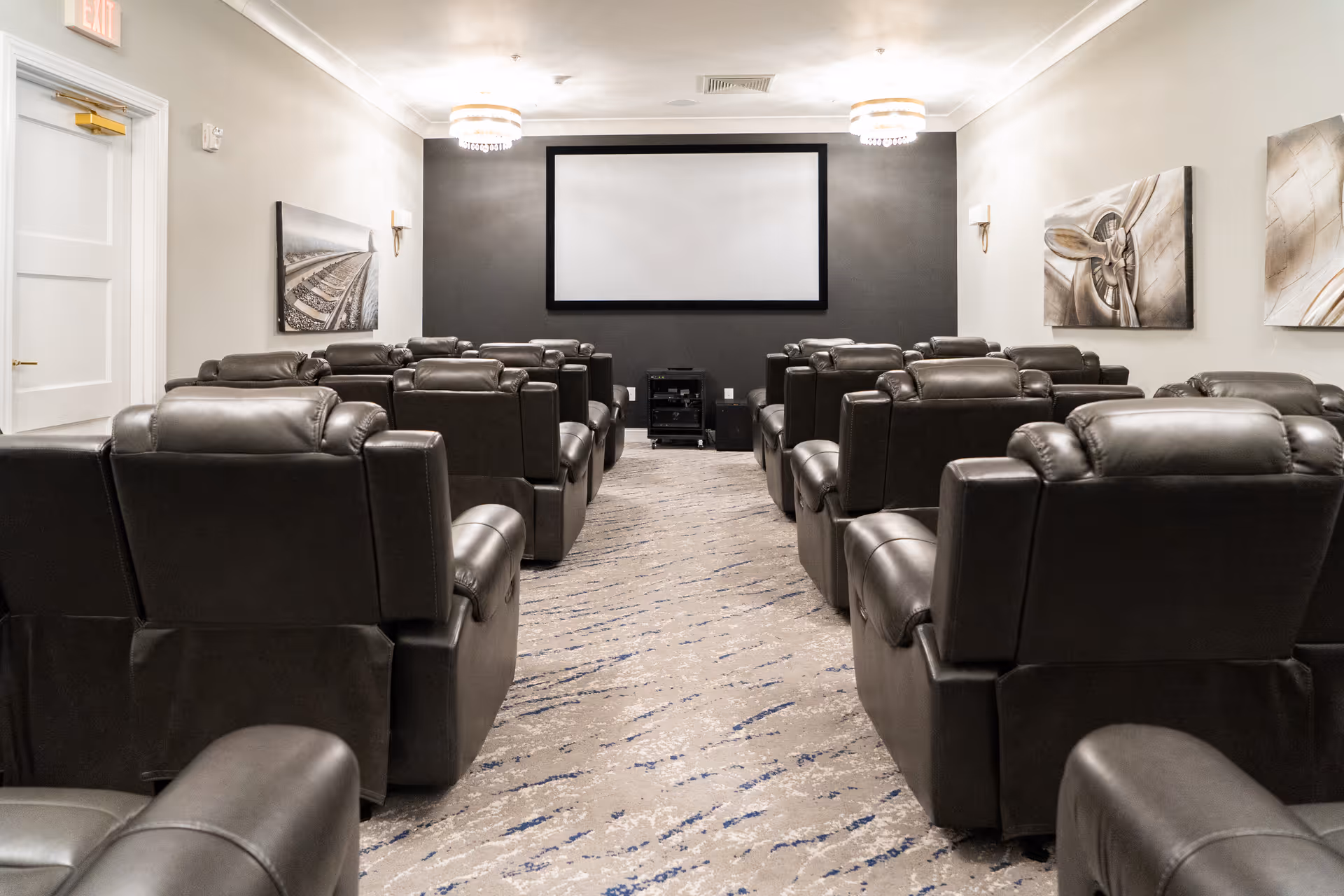 A small theater room with multiple rows of black leather recliner chairs facing a large blank projection screen on a dark gray wall. The room has light-colored walls with modern artwork and two ceiling light fixtures.