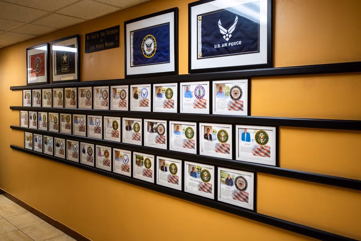 A hallway wall display featuring framed photos and descriptions of veterans, with military branch flags and a plaque that reads 'We Salute The Veterans of Lake Pointe Woods' above the frames.