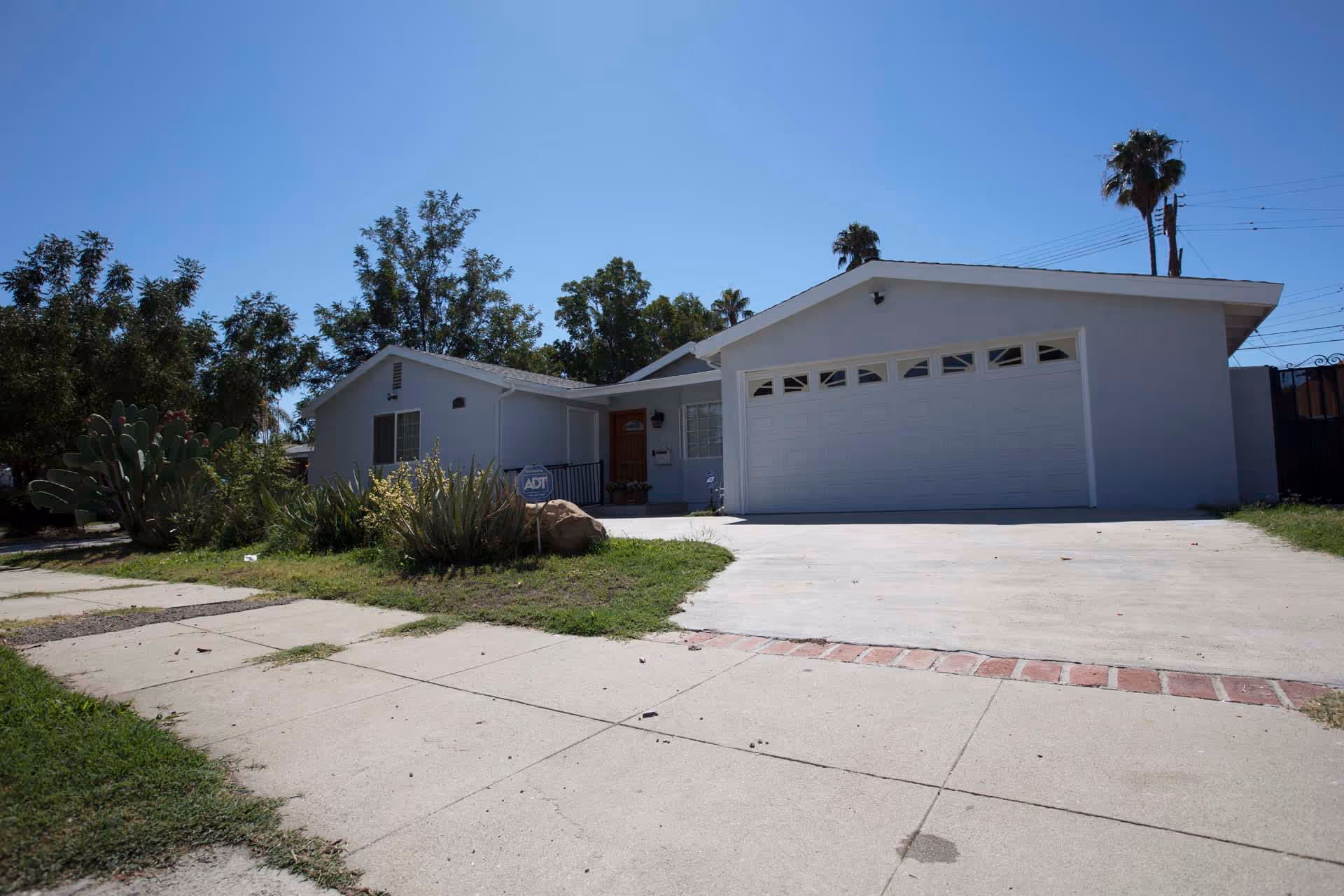 Front exterior of a single-story pale-gray house with a two-car garage, driveway, and small front yard landscaping under a clear blue sky.