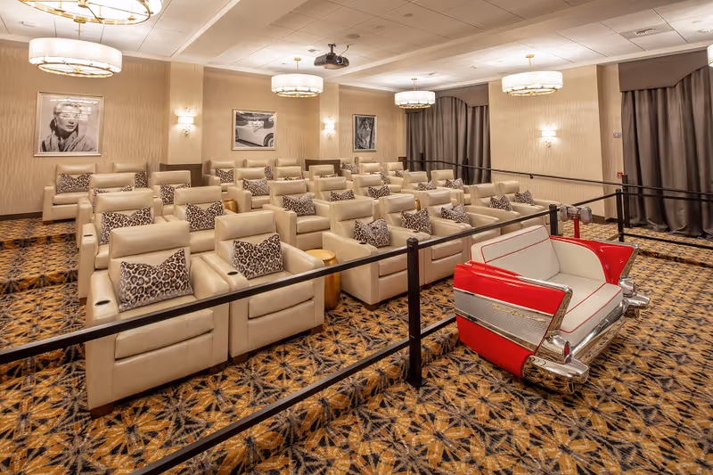 A cozy theater room with multiple rows of beige leather recliners, each with a leopard print pillow. The room features patterned carpet, framed black and white photos on the walls, and modern ceiling lights. A unique red and white retro car-themed loveseat is positioned at the front right side of the room.