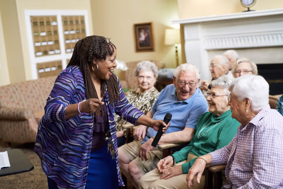A woman holding a microphone interacts with a group of elderly people seated in a cozy living room setting. The elderly individuals are smiling and appear engaged in the activity. The room features comfortable chairs, a fireplace, and warm lighting.