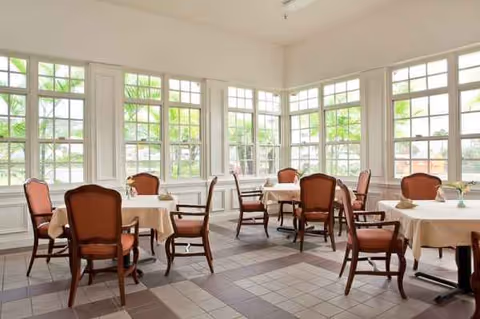 Bright dining room with round tables, light beige tablecloths, and wooden chairs.