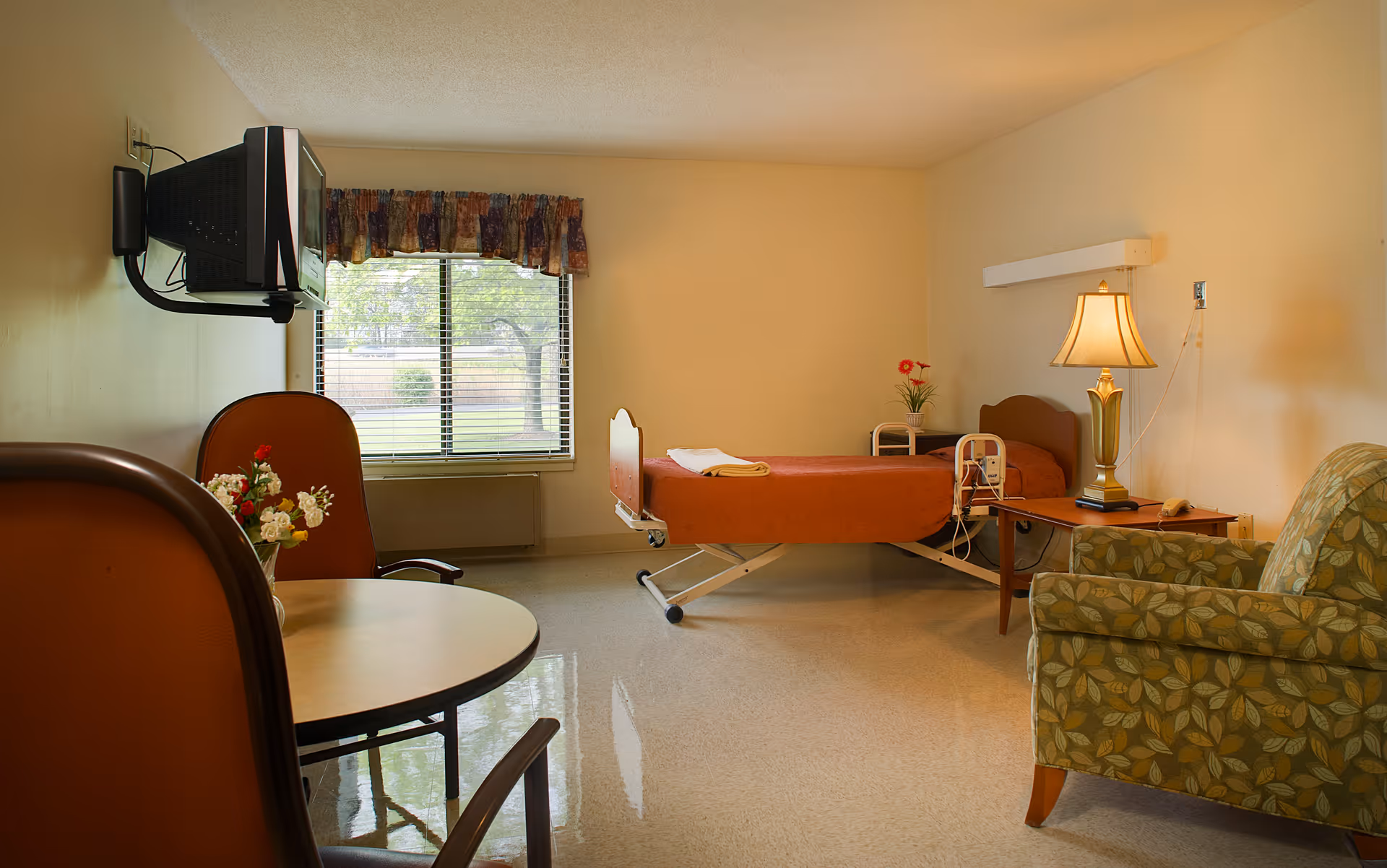 A simple senior living facility room with a single hospital-style bed covered in an orange blanket, a wooden side table with a lamp and a small plant, a green patterned armchair, a round table with two orange chairs, a wall-mounted TV, and a window with floral curtains showing greenery outside.