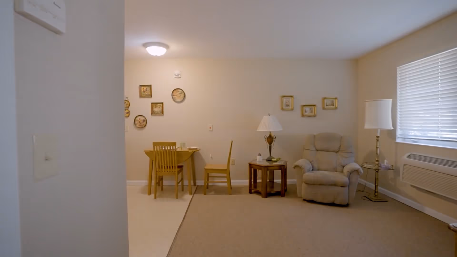 A cozy living area in an assisted living facility featuring a beige recliner chair, two side tables with lamps, a window with blinds, and a small dining table with two wooden chairs. The walls are decorated with framed pictures and the room has a warm, neutral color palette.
