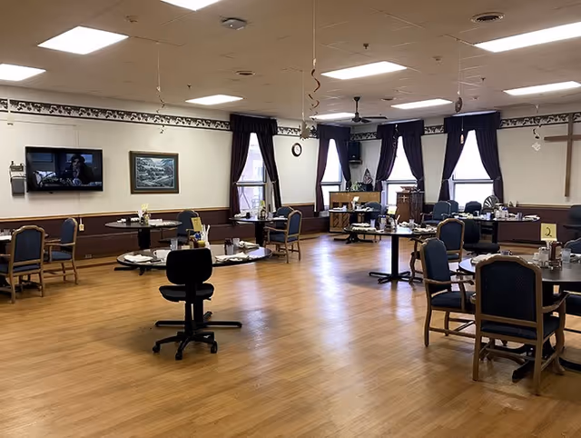 A spacious dining room with several round tables and chairs arranged around the room. The tables are set with plates, cups, and utensils. The room has wooden flooring, large windows with dark curtains, a wall-mounted TV, and a cross on the wall. The lighting is bright with ceiling lights.