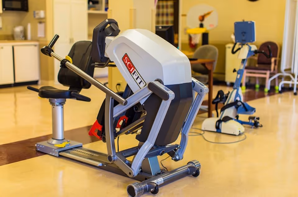 A rehabilitation exercise room featuring a SciFit exercise machine with a seat and handles, another piece of exercise equipment in the background, and a small basketball hoop on the wall.