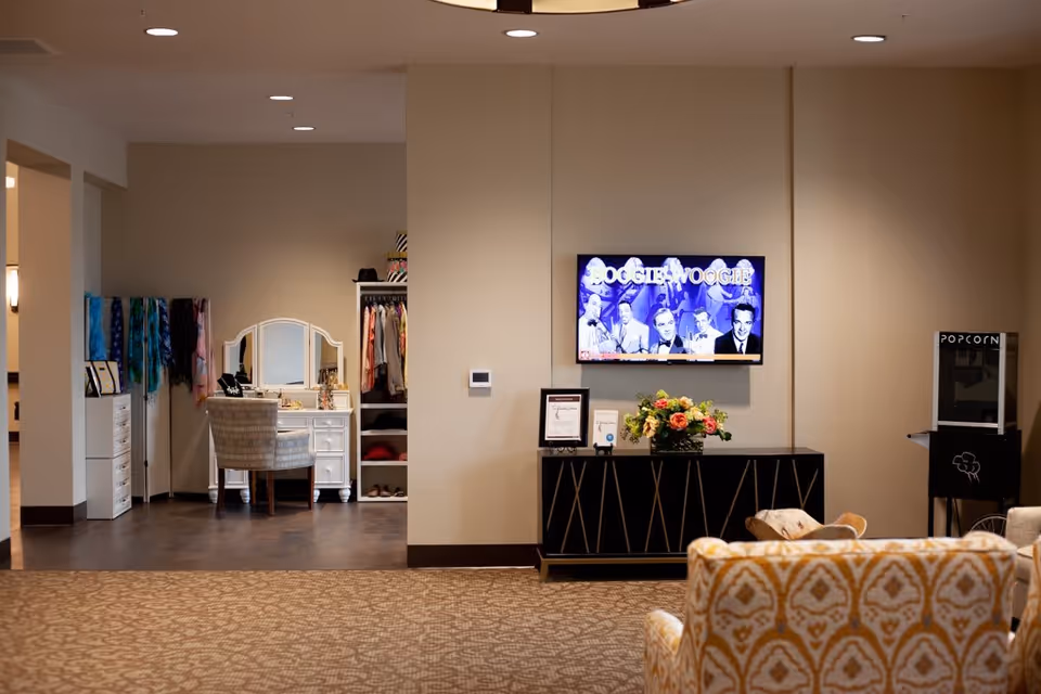 A cozy interior space featuring a patterned armchair in the foreground, a black cabinet with a flower arrangement and framed items, a wall-mounted TV displaying a Boogie Woogie show, and a popcorn machine to the right. To the left, there is a vanity table with a mirror and chair, scarves hanging on a rack, and an open closet with clothes and shoes.