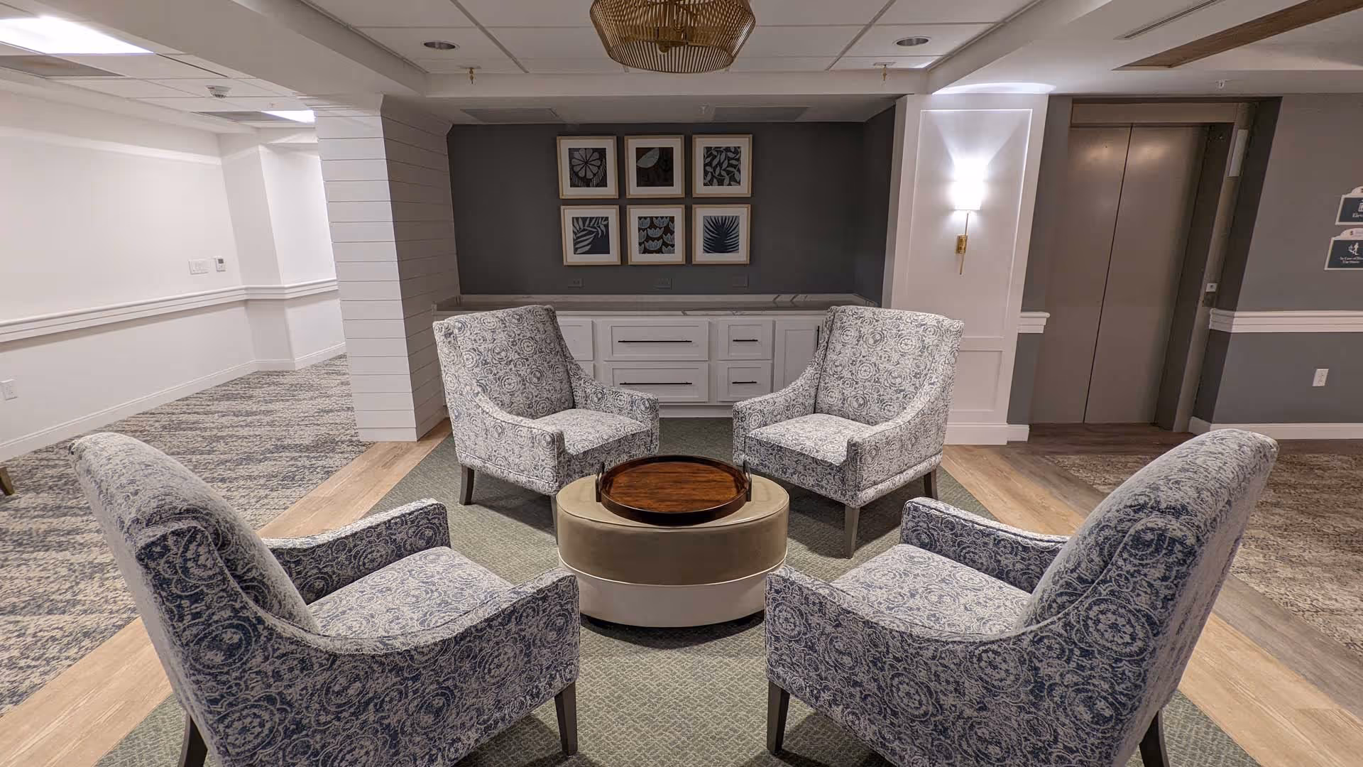 A cozy seating area in a senior living facility featuring four patterned armchairs arranged around a round ottoman with a wooden tray on top. The space has a neutral color palette with a gray accent wall adorned with six framed botanical prints. There is a white cabinet below the artwork and an elevator door to the right. The floor is a mix of carpet and wood, and the ceiling has recessed lighting and a decorative light fixture.