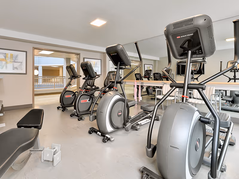 A senior living community fitness room with exercise equipment including stationary bikes, elliptical machines, a bench, and dumbbells. The room has a large mirror on one wall and framed artwork on the opposite wall. There is an open doorway leading to an outdoor or balcony area.