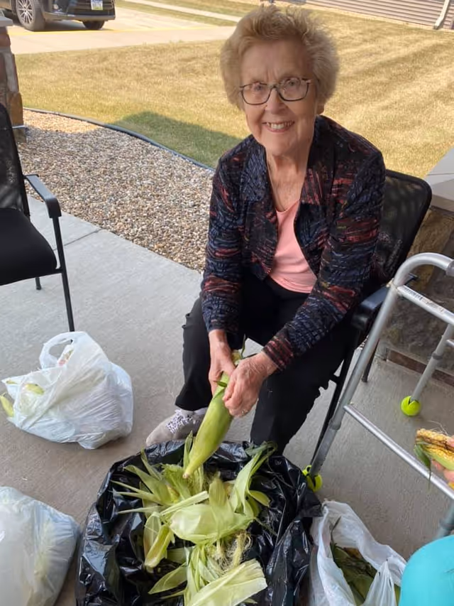 An elderly woman sitting outside on a chair, smiling while shucking corn. She is surrounded by bags and a black trash bag filled with corn husks. A walker with tennis balls on the legs is nearby, and a car is parked in the background on a driveway.