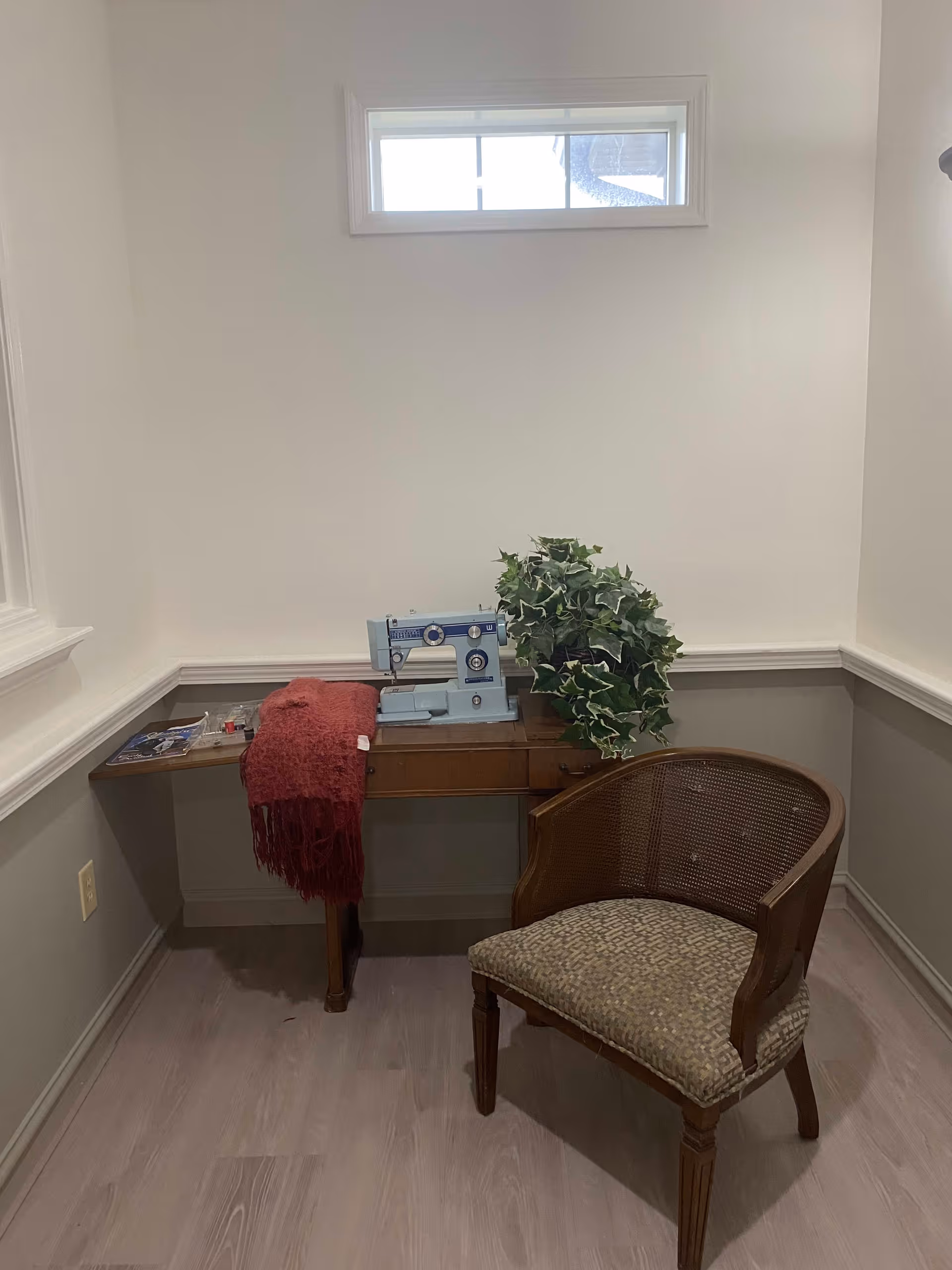 A small corner room with a wooden sewing table holding a vintage blue sewing machine, a red knitted scarf, and a green leafy plant. A cushioned wooden chair is placed in front of the table. The walls are painted white with a gray lower half and there is a small rectangular window near the ceiling.