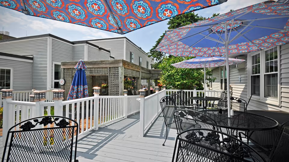 Outdoor patio area with black metal tables and chairs, each table shaded by large blue umbrellas with colorful patterns. The patio has a white railing and is adjacent to a light gray building with multiple windows. There is greenery and a wooden pergola in the background under a partly cloudy sky.