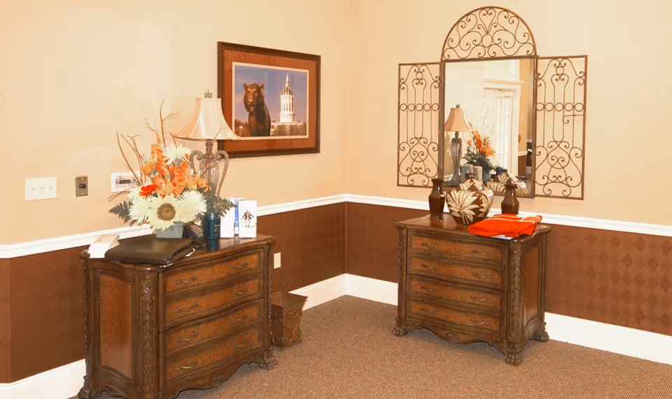 A corner of an interior room with two wooden dressers featuring ornate carvings. One dresser has a floral arrangement with orange and white flowers, a lamp, and some brochures. The other dresser has decorative vases and a folded orange cloth. Above the second dresser is a large mirror with a decorative wrought iron frame. On the wall above the first dresser is a framed picture of a tiger statue with a building in the background. The walls are painted beige with a brown wainscoting and white trim, and the floor is carpeted.