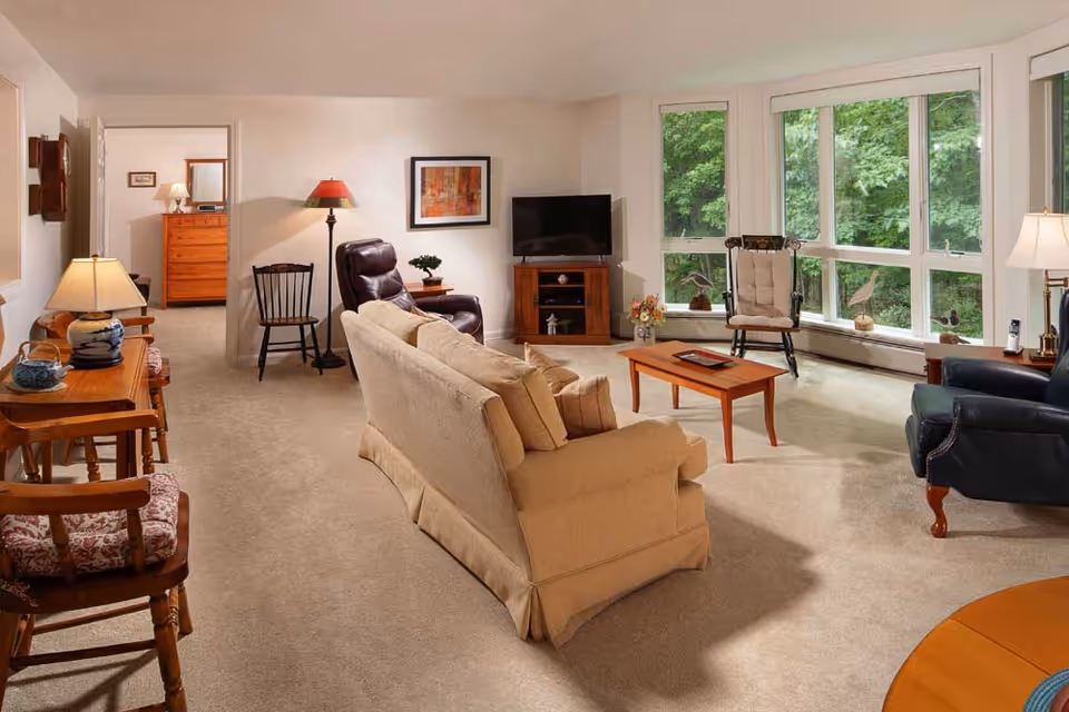 A cozy living room with beige carpet and walls, featuring a beige sofa with cushions, a wooden coffee table, a dark leather recliner, and a wooden TV stand with a flat-screen TV. Large windows on one side offer a view of green trees outside. Additional wooden chairs and lamps are placed around the room, and a doorway leads to a bedroom with a wooden dresser and lamp visible.
