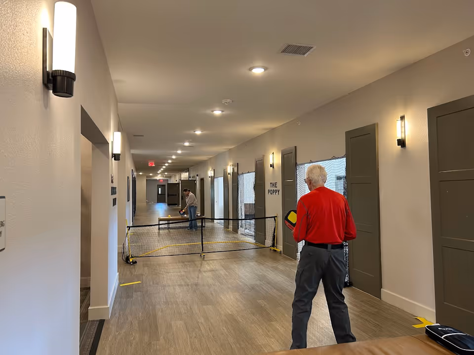 Two elderly men playing pickleball in a long, well-lit hallway with wood flooring and beige walls. One man in a red shirt is in the foreground holding a paddle, while the other man is in the background preparing to hit the ball. A small net is set up across the hallway, and doors line the walls on both sides.