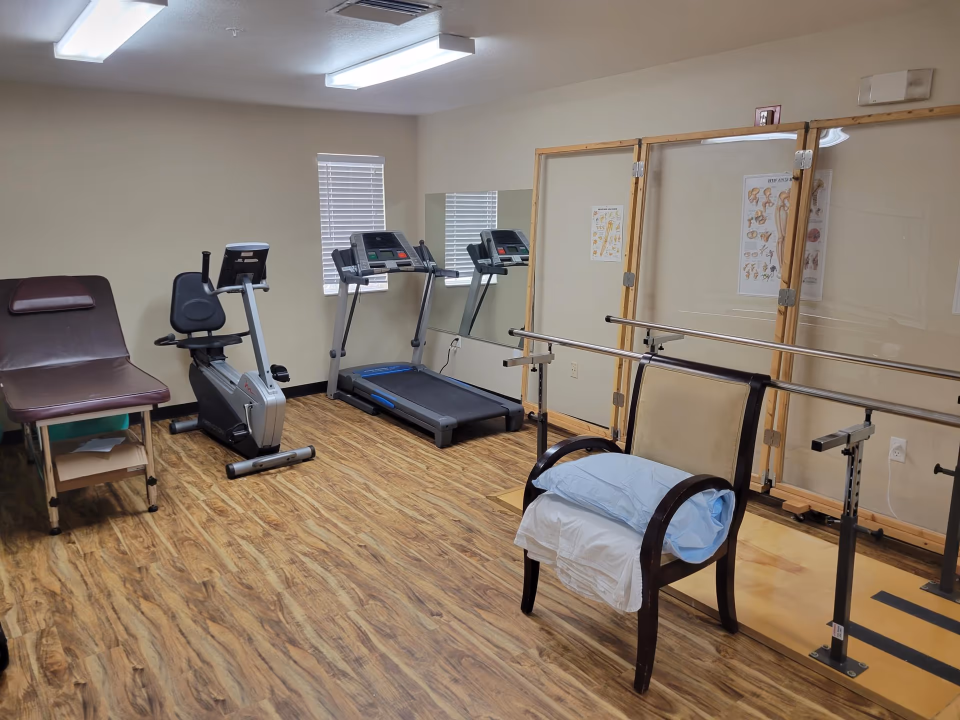 A rehabilitation or physical therapy room with exercise equipment including a recumbent bike, treadmill, parallel bars for walking support, a padded examination table, and a chair with pillows. The room has wood-patterned flooring, beige walls, two windows with blinds, and anatomical posters on the wall.