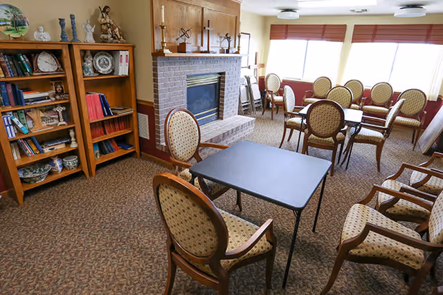 A cozy common area with patterned carpet, several upholstered chairs around tables, a brick fireplace with a wooden mantel, and a wooden bookshelf filled with books and decorative items. Large windows with blinds allow natural light to fill the room.