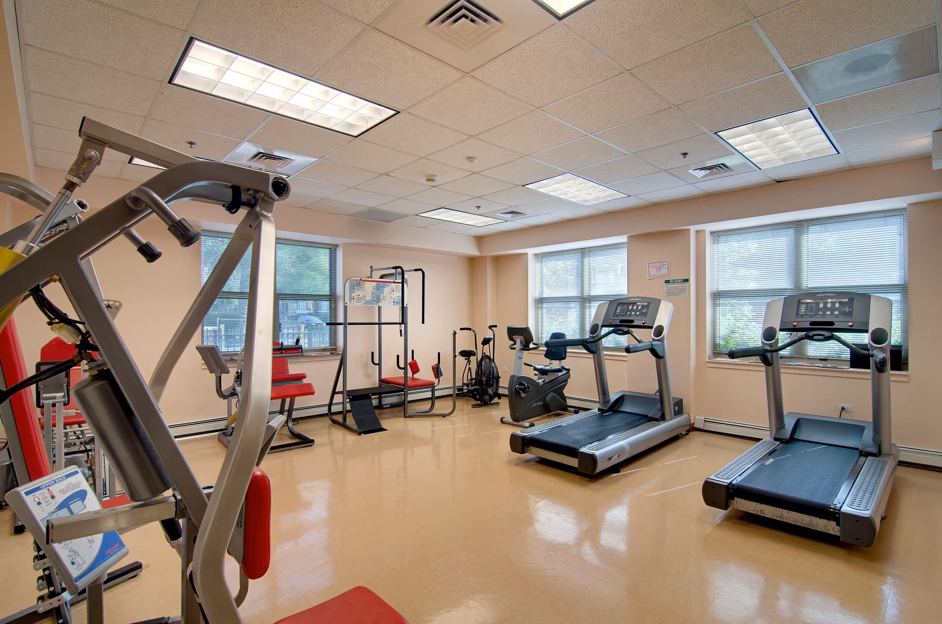 A well-lit fitness room with exercise equipment including two treadmills, stationary bikes, and various strength training machines. The room has large windows with blinds, beige walls, and a polished floor.