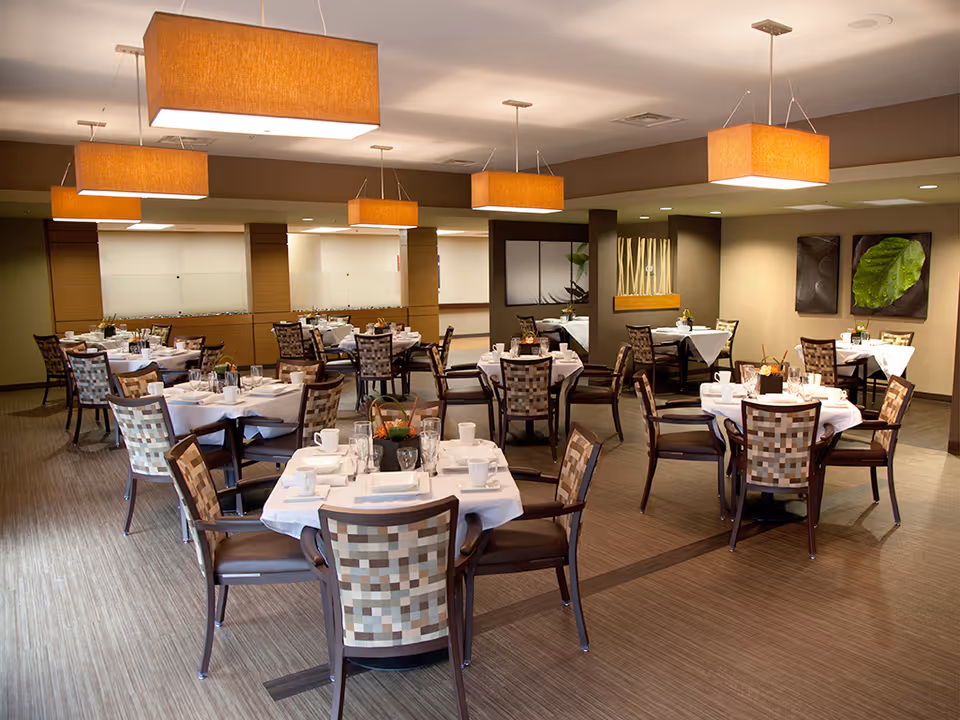 A well-lit dining room with multiple round tables covered with white tablecloths, each set with cups, glasses, napkins, and small decorative centerpieces. The room features modern pendant lights with rectangular orange shades hanging from the ceiling, patterned chairs, and neutral-toned walls with framed leaf artwork.