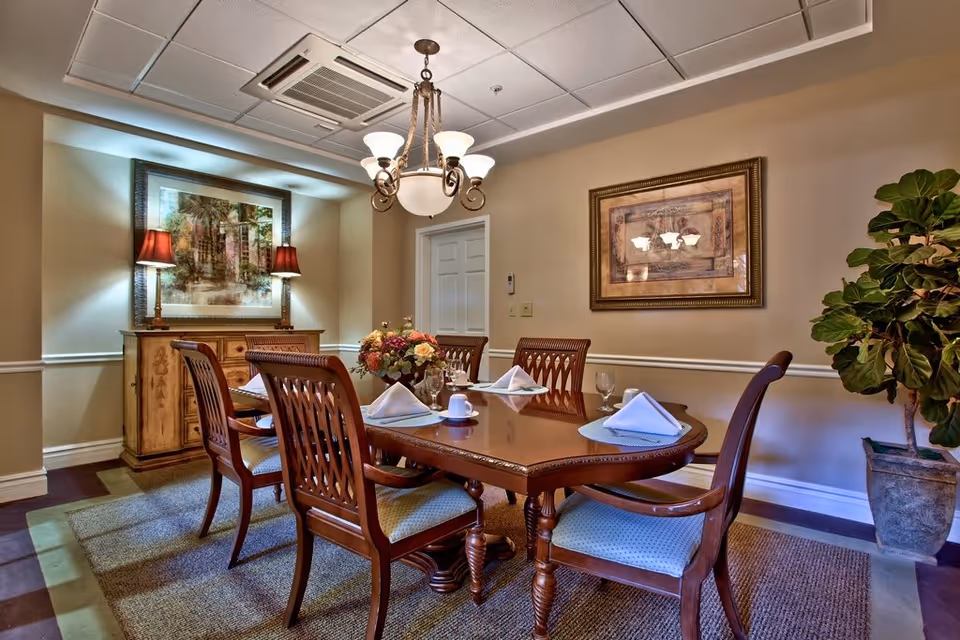 A formal dining room with a polished wooden table set for six, featuring folded white napkins, glassware, and a floral centerpiece. The room has beige walls adorned with framed artwork, two table lamps on a sideboard, a chandelier overhead, and a large potted plant in the corner.