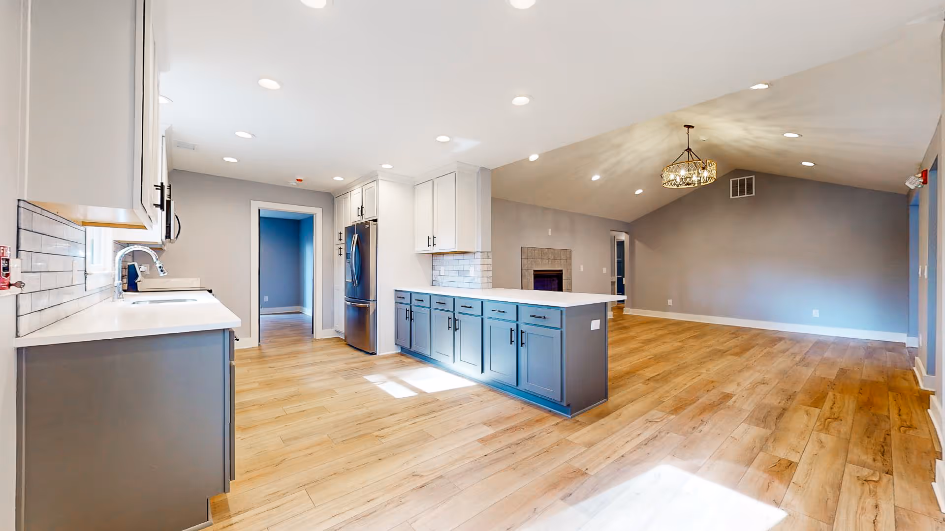 Bright and spacious open-concept interior featuring a modern kitchen with white countertops, gray lower cabinets, white upper cabinets, stainless steel refrigerator, and a kitchen island. The kitchen opens into a large living area with wood flooring, a fireplace, vaulted ceiling, and a decorative chandelier.