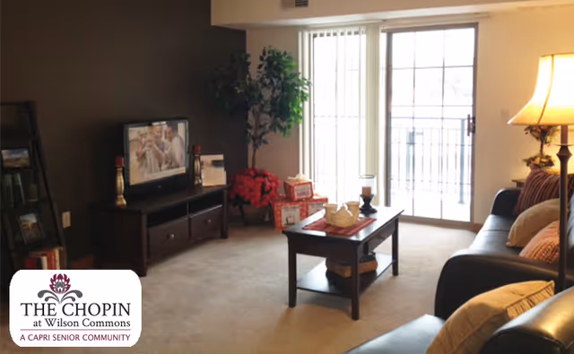 A cozy living room with a dark brown accent wall, a flat-screen TV on a wooden TV stand, a coffee table with decorative items, a black leather sofa with cushions, a floor lamp, and a sliding glass door leading to a balcony. There are also some plants and wrapped gifts near the TV stand.