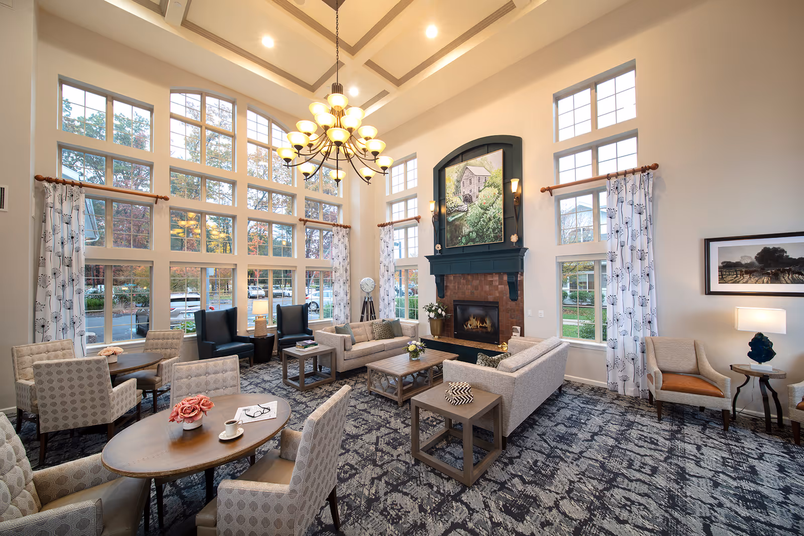 Bright, spacious senior living common lounge with tall multi-pane windows, chandelier, fireplace, sofas and multiple seating tables.