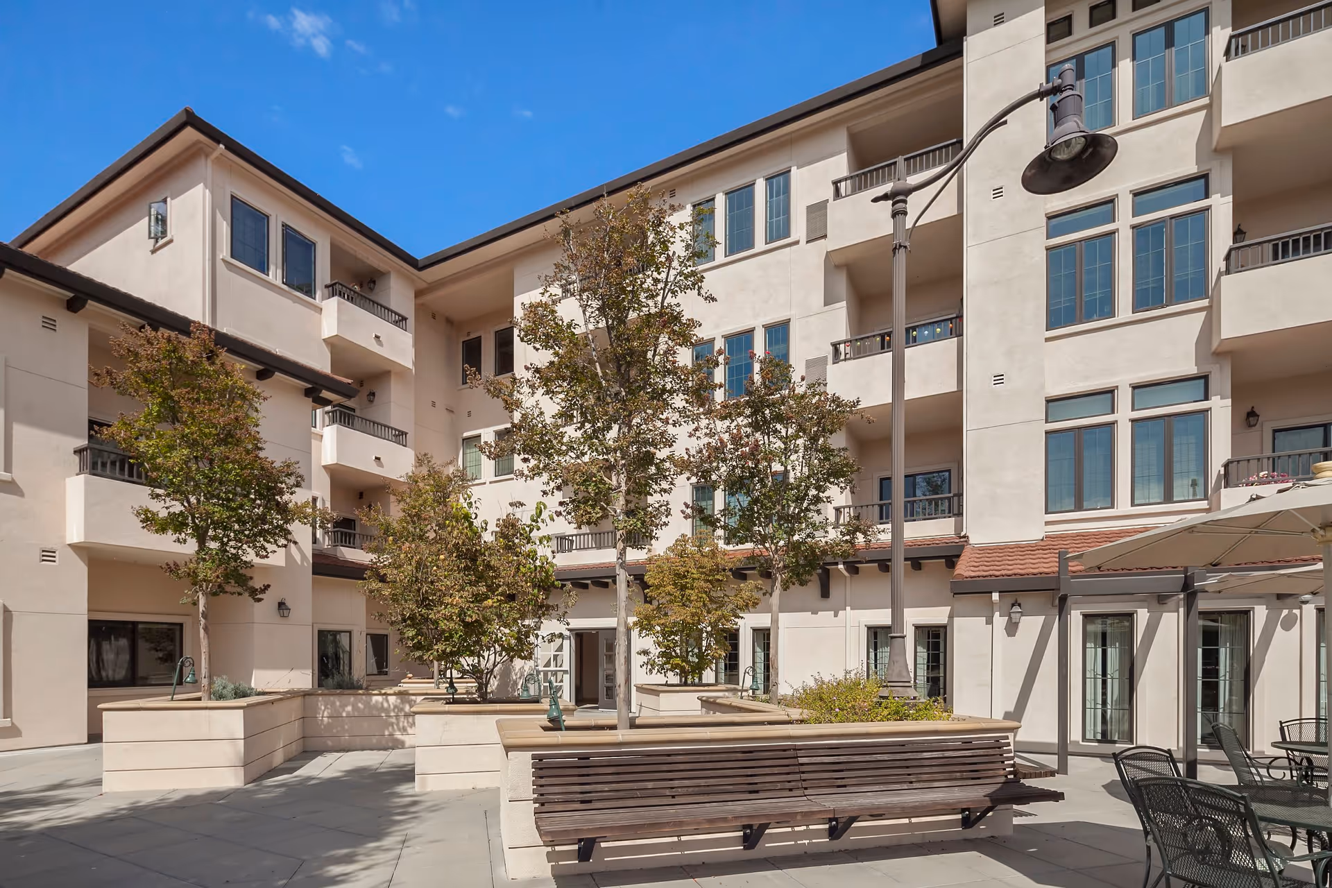 Outdoor courtyard area of a senior living facility with several trees planted in raised planters, a wooden bench, metal chairs and tables under a large umbrella, and a multi-story building with balconies and large windows in the background under a clear blue sky.