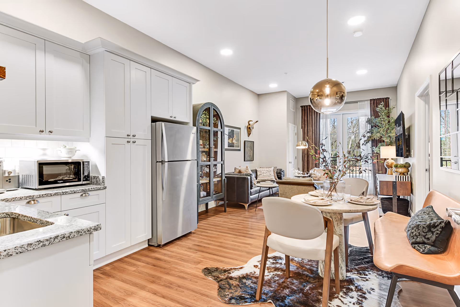 Bright and modern senior living space featuring a kitchen with white cabinets, granite countertops, and stainless steel refrigerator. Adjacent dining area has a round table set with plates and napkins, surrounded by chairs and a bench. The living area includes a leather sofa, armchair, decorative cabinet, and large windows with curtains letting in natural light. Hardwood floors and contemporary lighting fixtures complete the cozy and stylish interior.