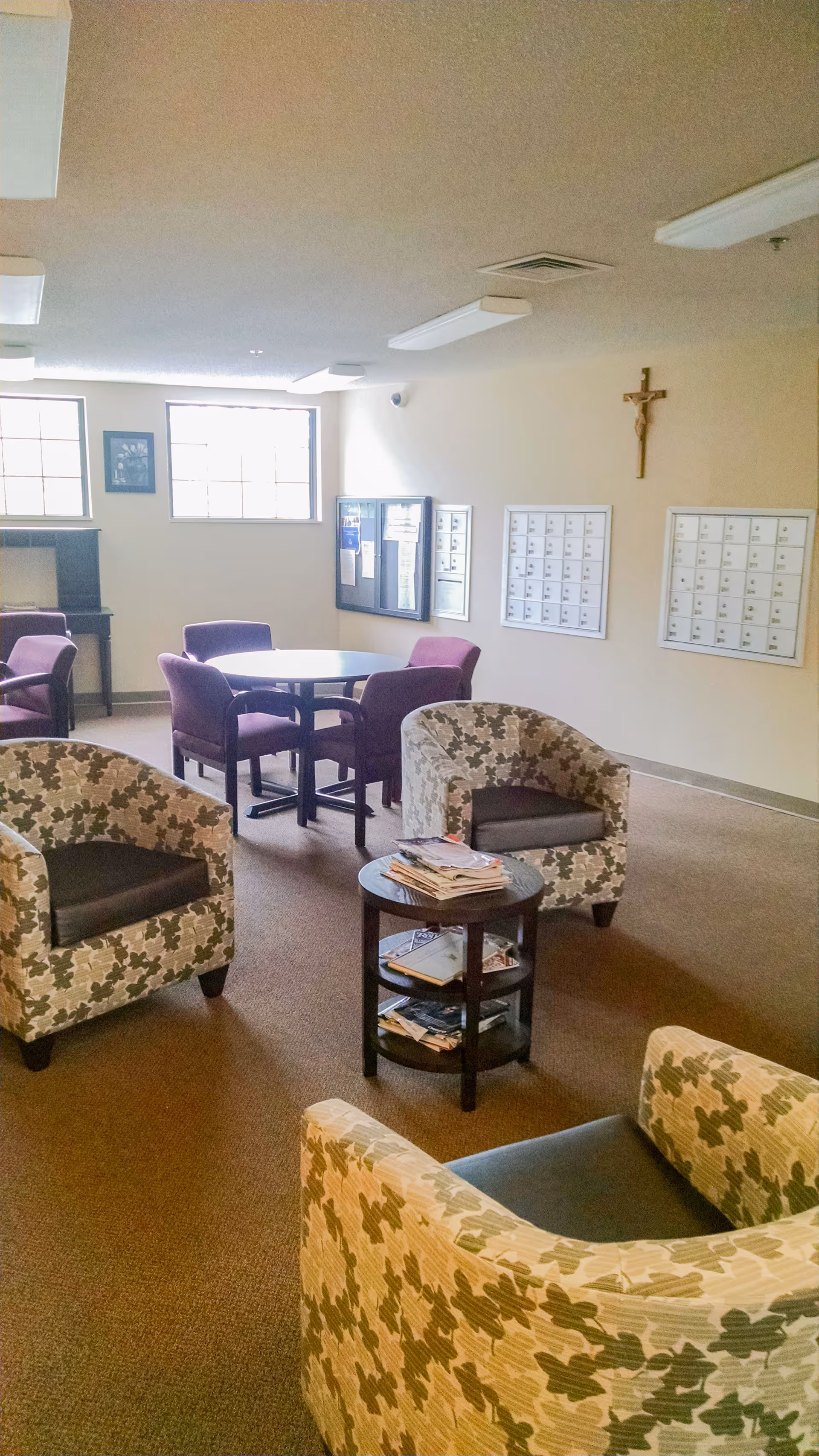 A common area in a senior living facility with patterned armchairs, a round table with purple chairs, a small round table with magazines, windows letting in natural light, mailboxes on the wall, a crucifix, and a bulletin board.