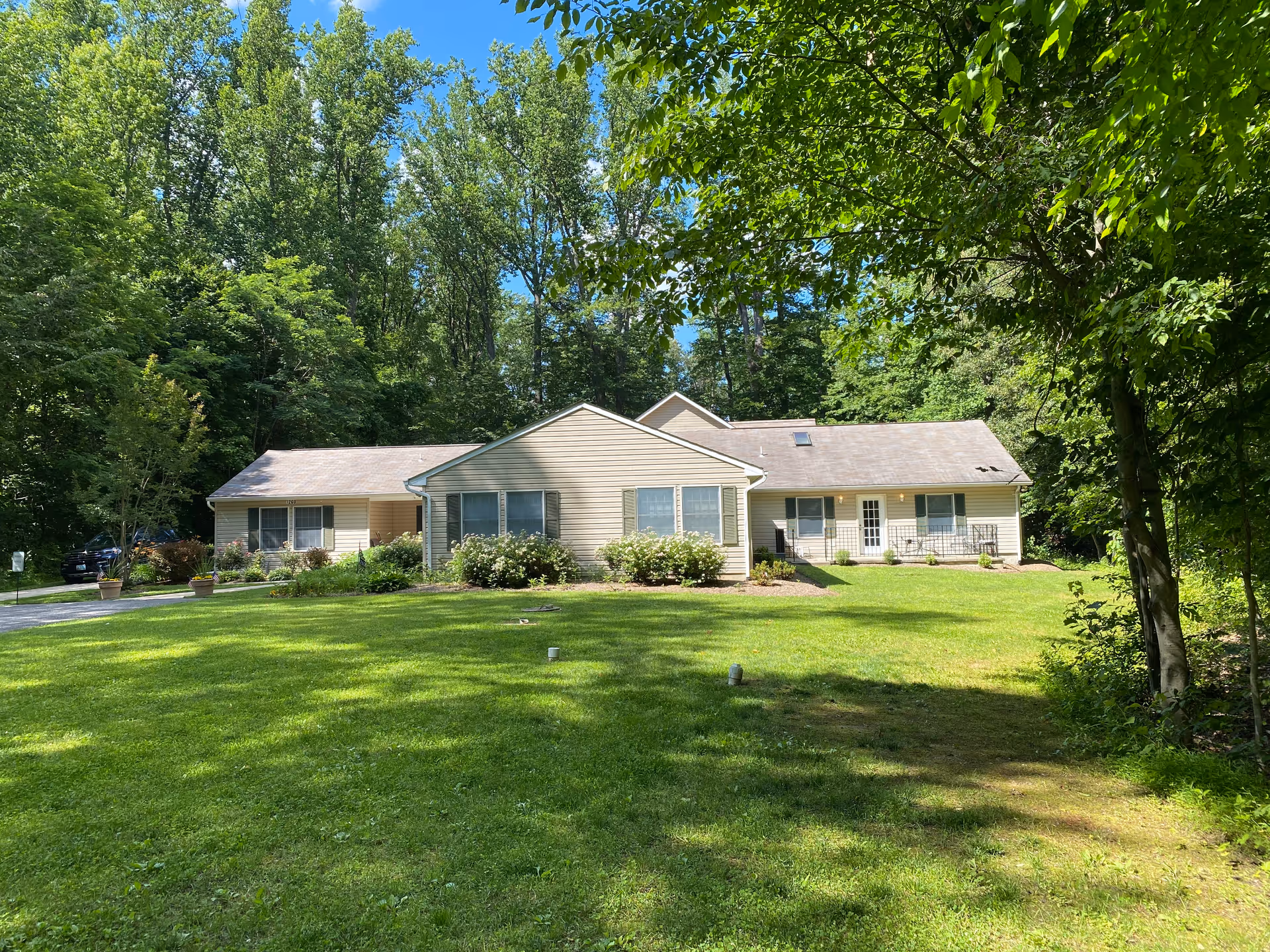 Single-story beige building with multiple windows and a door, surrounded by a large green lawn and tall trees under a blue sky.
