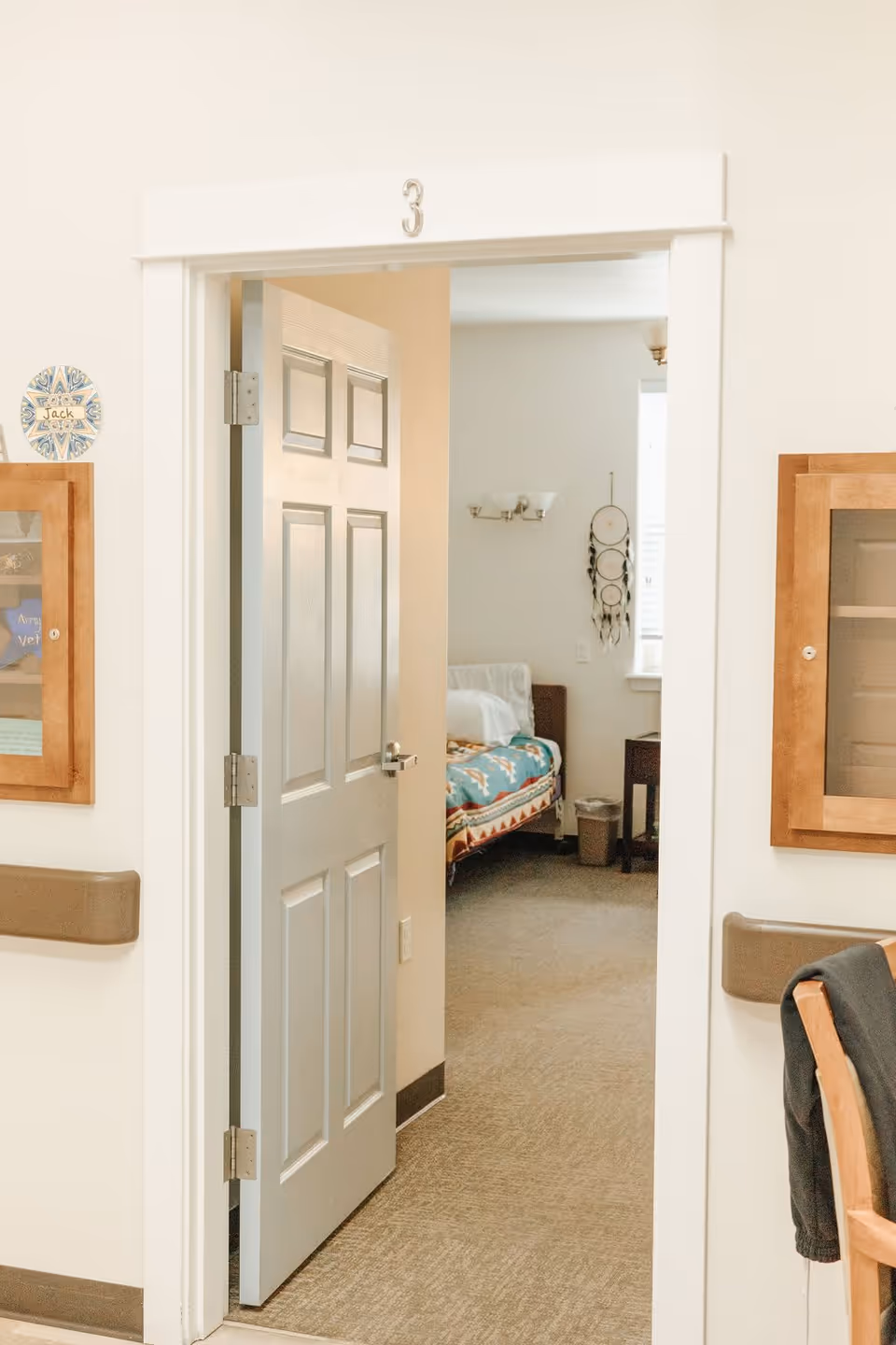 View through an open door labeled with the number 3 into a bedroom with a bed covered by a colorful blanket, a nightstand, a window with blinds, and a dreamcatcher hanging on the wall.