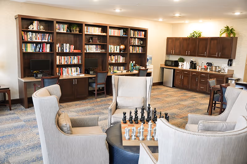 A cozy common room with wingback chairs around a table with a chess set, bookshelves with computers, and a kitchenette in the background.