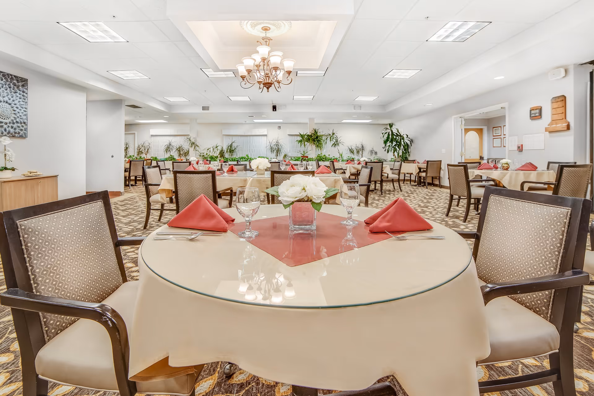 A spacious dining room with multiple round tables covered with beige tablecloths and glass tops. Each table is set with red folded napkins, clear drinking glasses, and silverware. The room is well-lit with ceiling lights and a central chandelier. There are several chairs around each table and green plants along the back wall, creating a welcoming atmosphere.