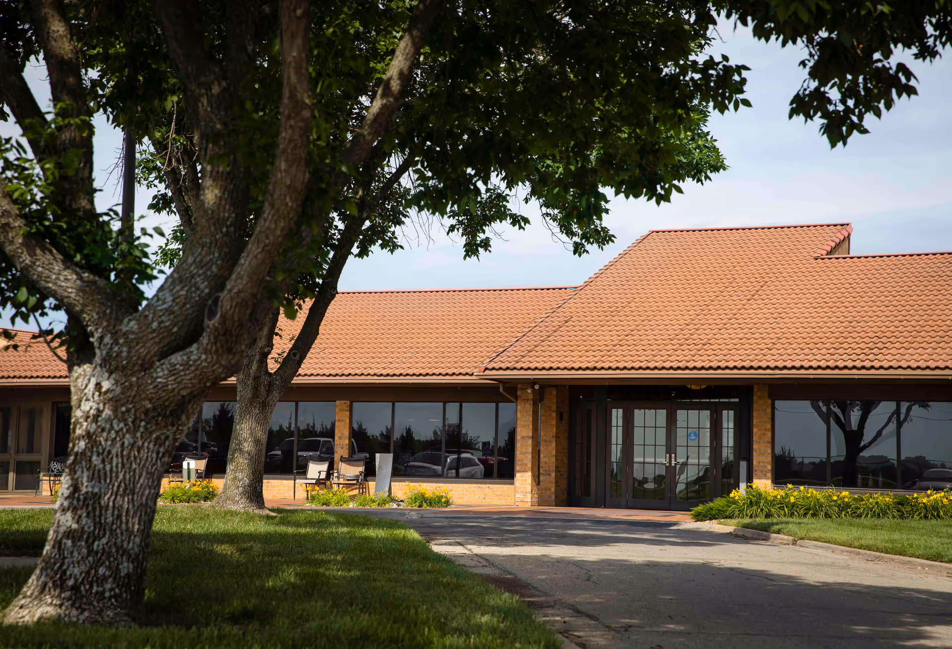 Front exterior view of a single-story building with a red tiled roof and large windows. There are trees and green grass in the foreground, and a paved driveway leading to the entrance with glass double doors.